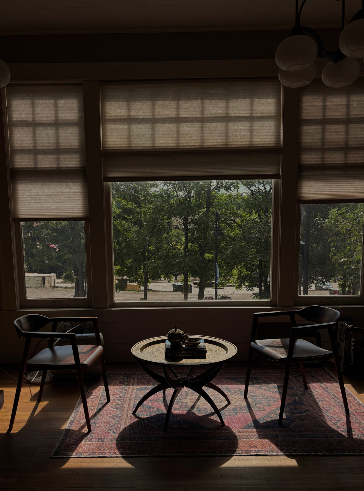 A dimly lit interior room with large windows showing a tree-lined street outside. Two dark chairs sit on either side of a small round table on a patterned rug, with hanging pendant lights visible at the top.